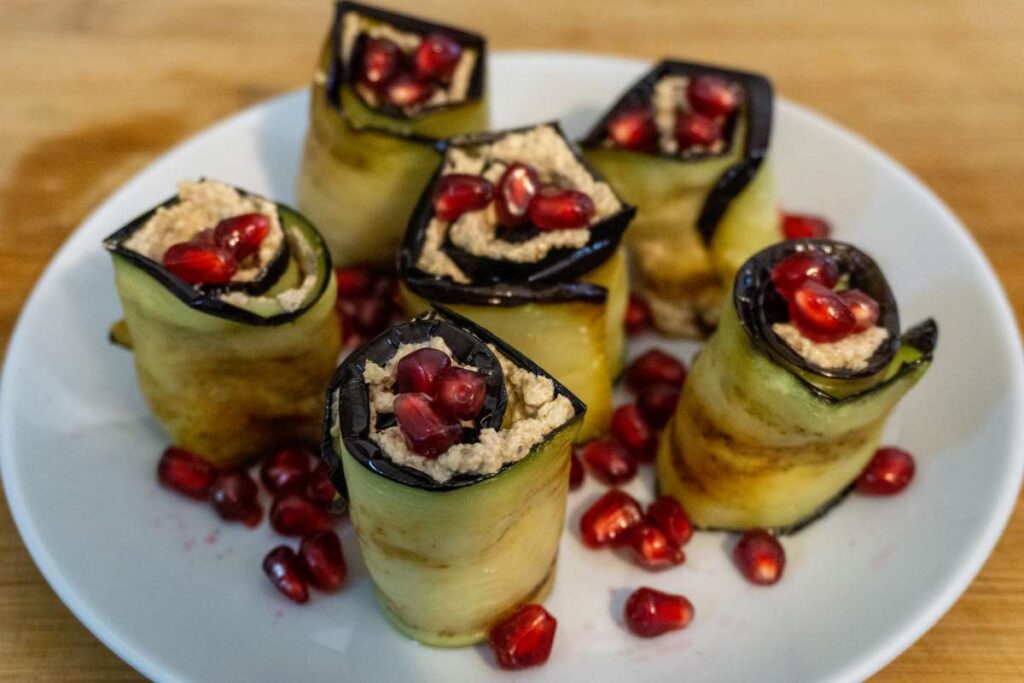 Georgian Eggplant Rolls by No Frills Kitchen, 6 rolls of egg plant on a white plate, sprinkled with pomegranate seeds