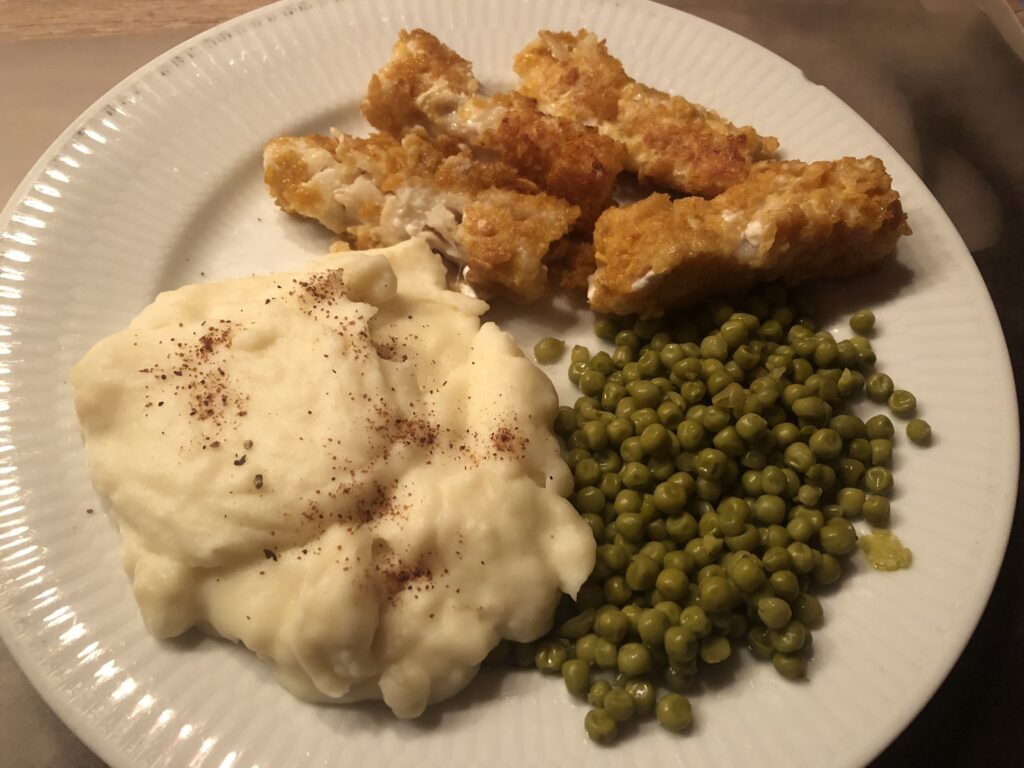 Fishsticks, peas and mashed potatoes, served on a white plate. Fishsticks on top, below on the right the peas on the left the mashed potatoes sprinkled with nutmeg